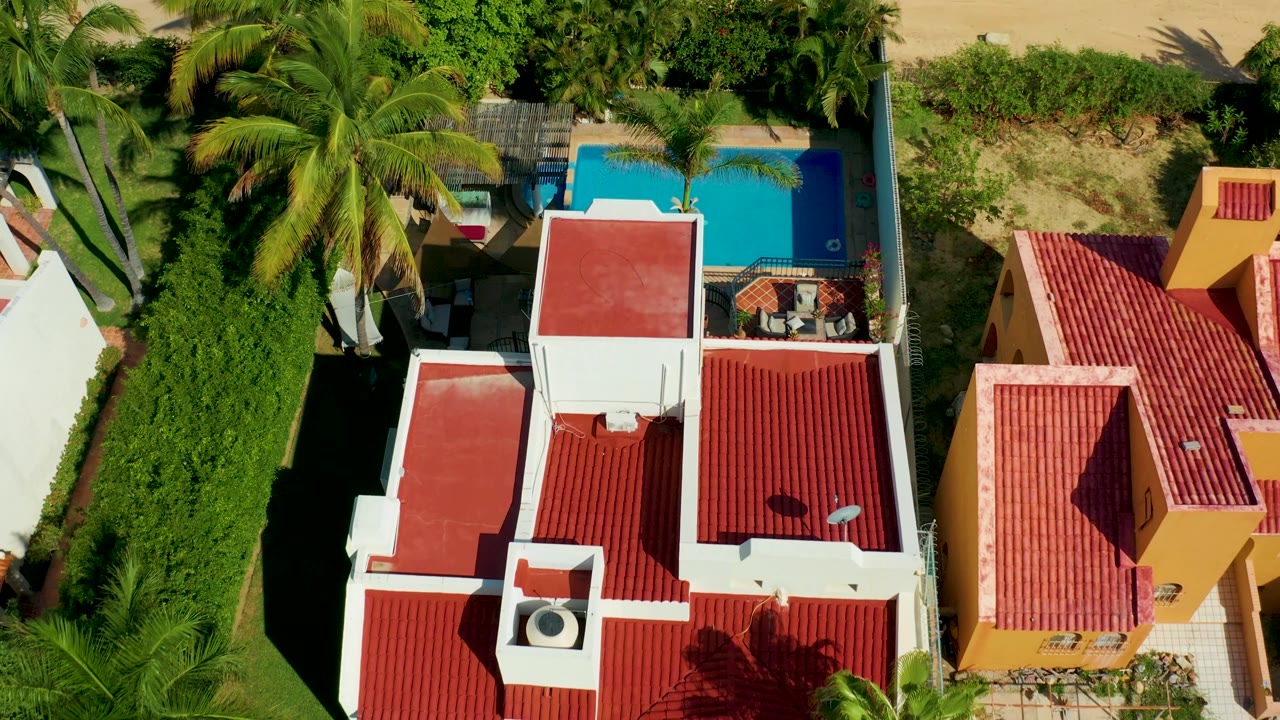 Overhead aerial frame in San Jose del Cabo showing white buildings with red tiled roofs, a blue pool near palms and hedges, and no visible people.