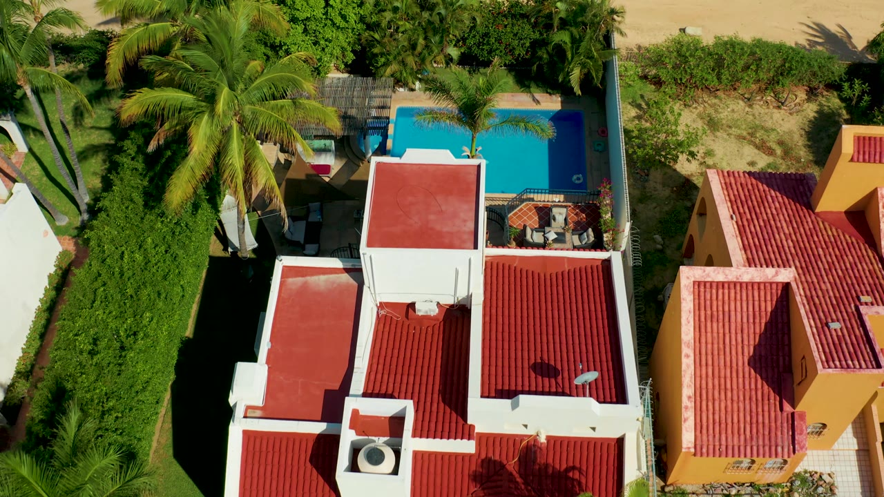 Top-down San Jose del Cabo frame showing a white residential block with red roofs, the blue swimming pool at upper right, palms and hedges, and no visible people.