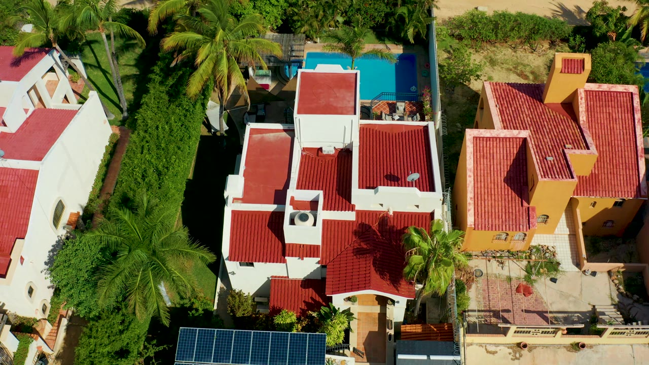 Overhead drone frame in San Jose Del Cabo showing white and yellow villas with red tiled roofs, tall palms, a blue pool, roof solar panels at the lower edge, and no visible people.