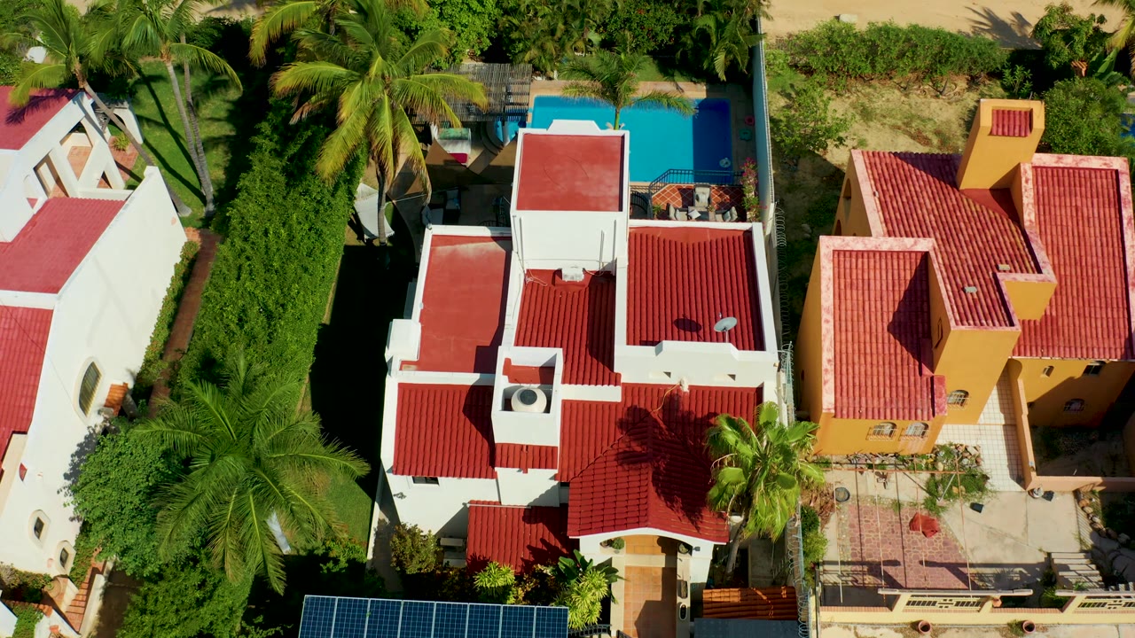 Top-down San Jose Del Cabo frame showing the white villa sliding toward the left side, the yellow building on the right, a blue pool at the upper edge, thick palms, and no visible people.