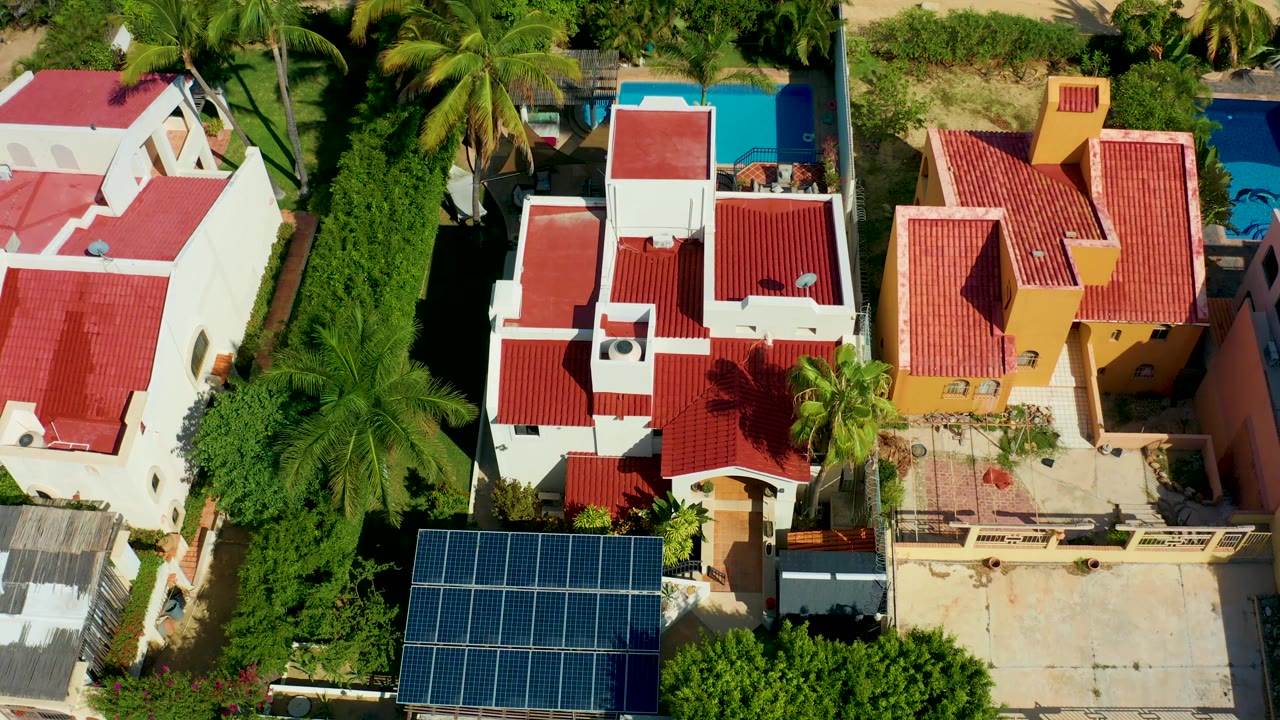 Aerial residential rooftops with red tiles, a blue pool, palms, and dark shadows.