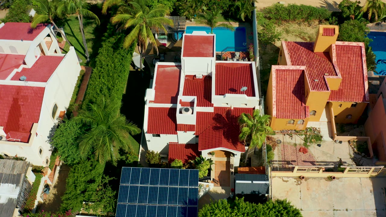 Residential aerial view with red roofs, palms, solar panels, pool, and courtyard shadows.
