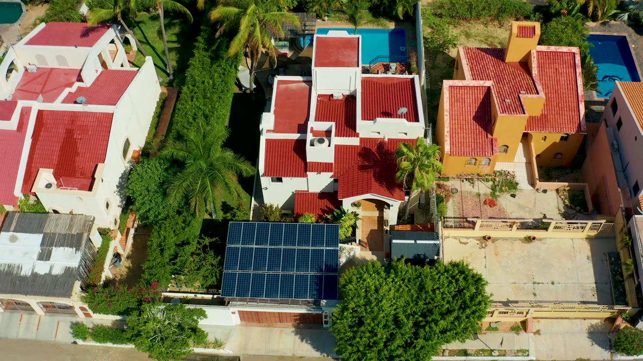 Overhead drone frame in San Jose del Cabo showing red-tiled residential rooftops, a solar-panel array, palms, greenery, and a blue pool with no visible people.