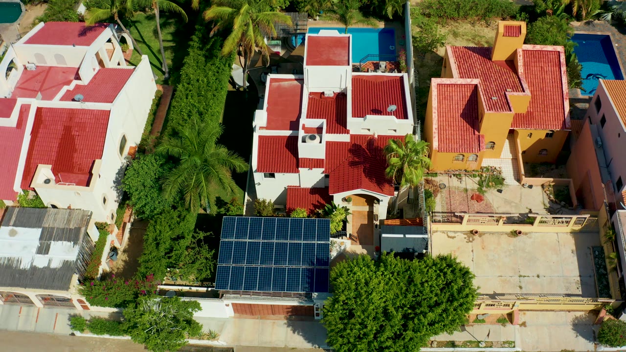 A second overhead view of San Jose del Cabo homes shows the solar-panel roof lower in frame, red roofs, palms, a yellow house, and pools with no visible people.