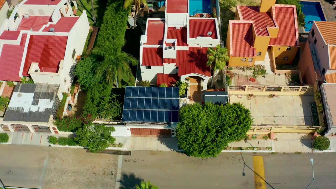 Overhead drone frame of San Jose Del Cabo homes with red roofs, a blue solar panel array, green trees, and a paved street at the bottom, with no visible people.