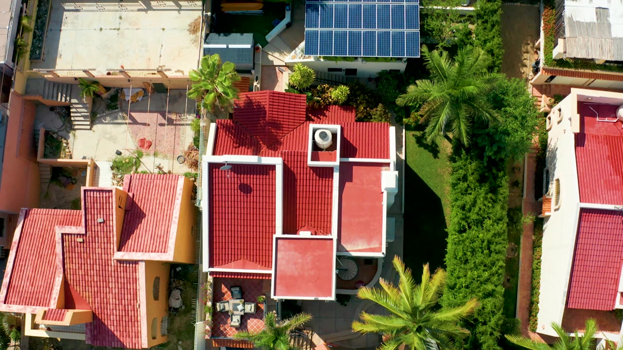 Overhead San Jose Del Cabo residential view with the red roofs shifted across frame beside a flat roof, blue solar panels, palms, trimmed greenery, and no visible people.