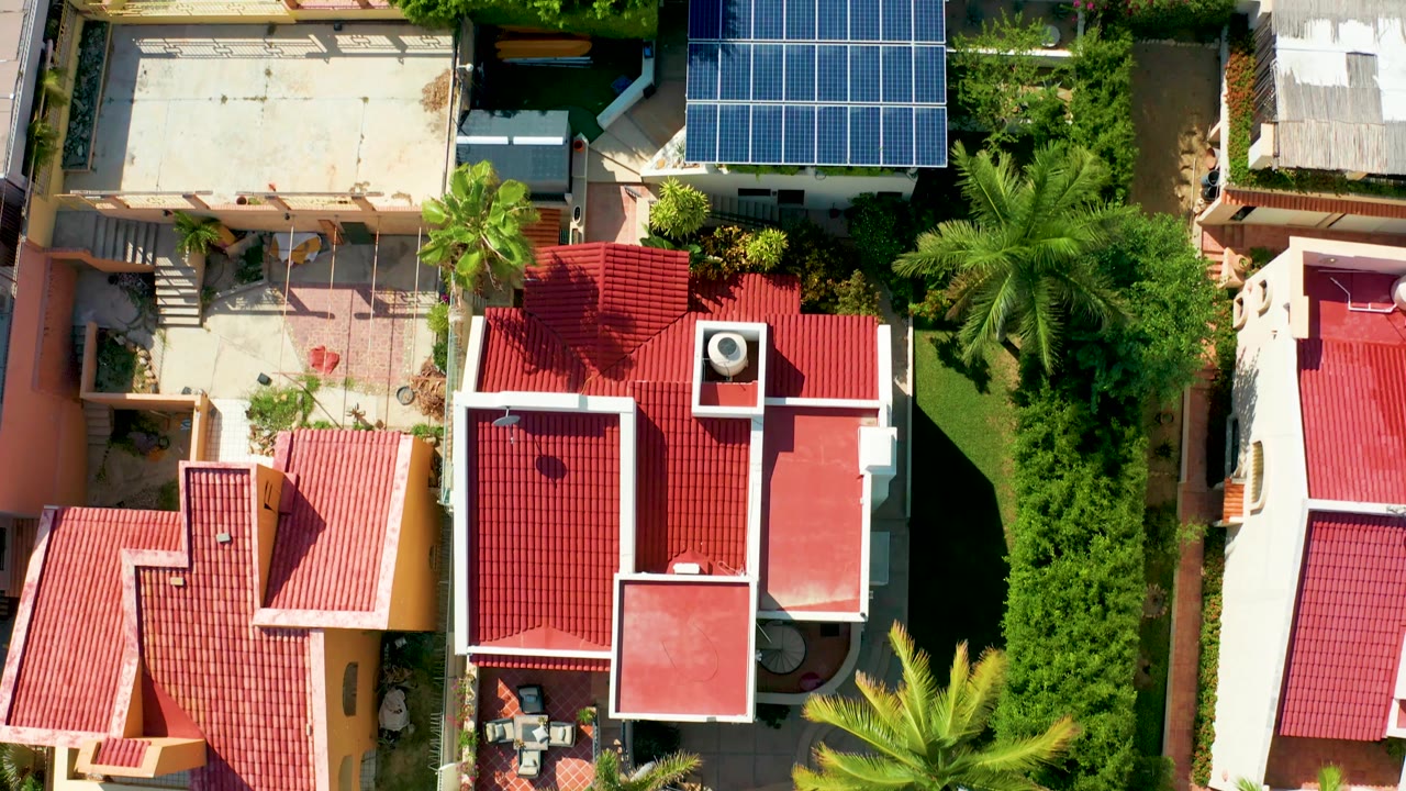 Top-down frame in San Jose Del Cabo showing red tiled residential roofs, a bright blue solar panel grid, palms casting shadows, green planting, and no visible people.
