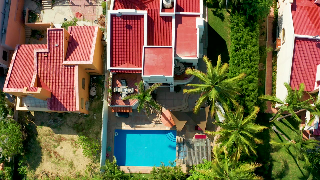 Overhead drone frame in San Jose Del Cabo showing red tiled residential roofs, a bright blue pool at lower left, palm trees, patios, grass, and no visible people.