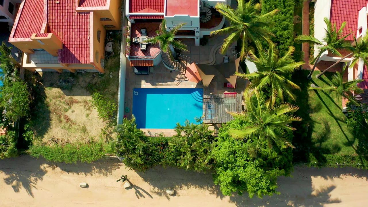 Overhead San Jose Del Cabo frame showing a red-roofed residence, bright blue pool, palms, garden greenery, sandy ground, and no visible people at the start of the clip.