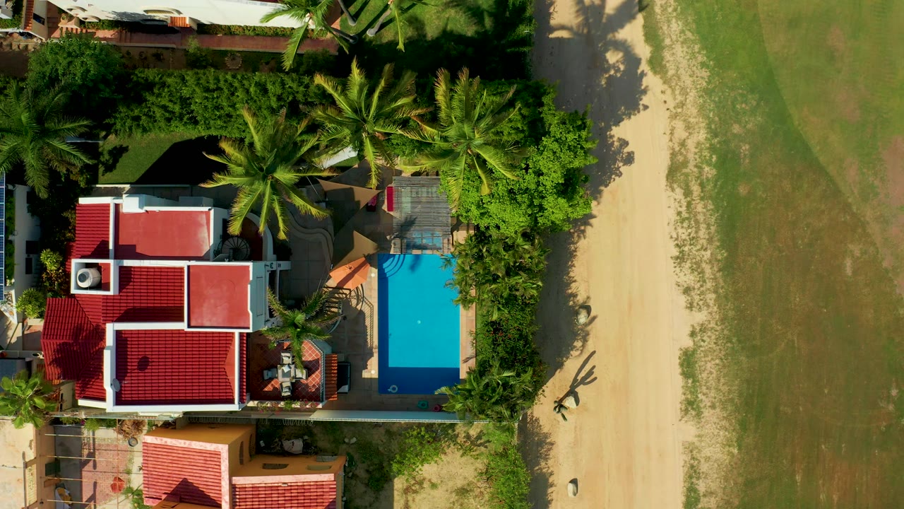 Top-down aerial of house with red roofs, pool, palms, sand path, and field.