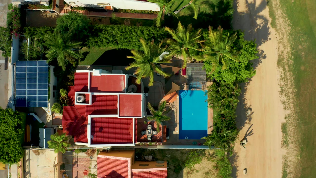 Top-down residential property with red roofs, solar panels, pool, trees, sandy path, and grass.