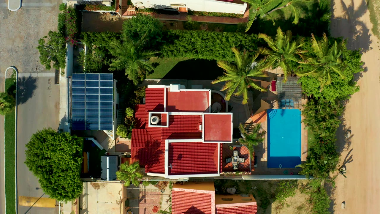 Top-down aerial frame in San Jose del Cabo showing a red-roofed residence, solar panels, dense trees, a blue pool, roads on both sides, and a small person visible on the roof.