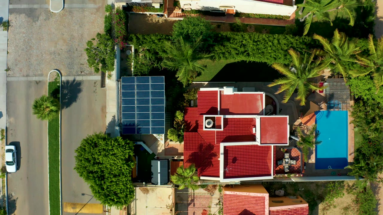A San Jose Del Cabo residential block appears from directly above, with the white car slightly farther down the left road beside solar panels, red roofs, a blue pool, and no visible people.