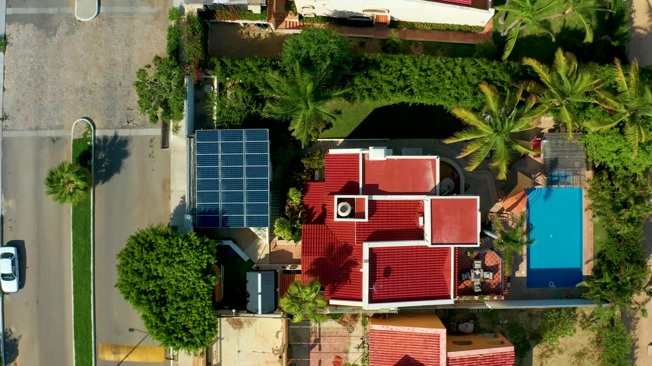 The overhead drone view holds on the San Jose Del Cabo home as the white car continues along the left-side street, with red tile roofs, solar panels, a bright blue pool, and no visible people.