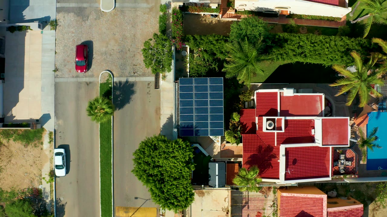 Red car moves down a residential street beside homes with solar panels and palms.