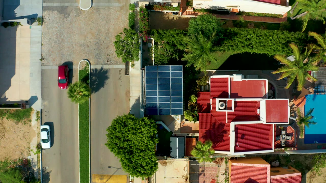 At 0:02, the red car continues along the residential street in San Jose del Cabo, with the parked white car below it, blue solar panels, red tile roofs, dense trees, and no visible people.