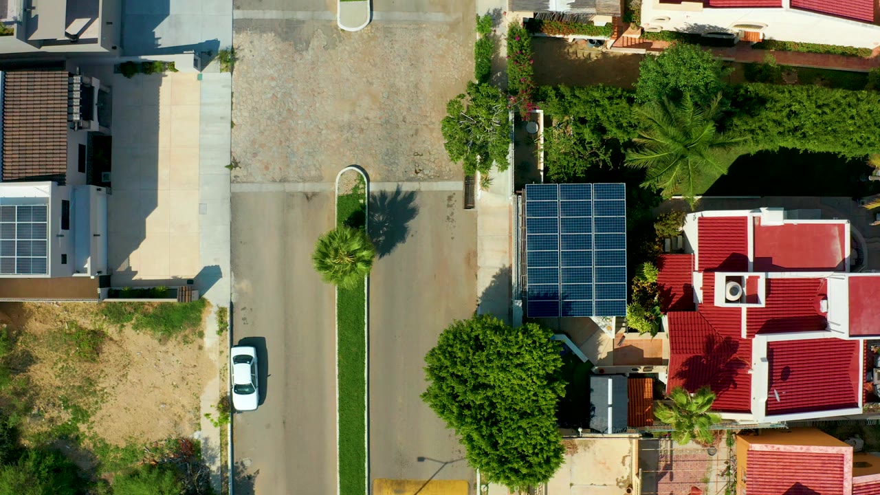 Top-down view in San Jose del Cabo of a residential street with a white car parked at the curb, solar panels beside a red-roofed house, palms, grass, dirt lots, and no visible people.