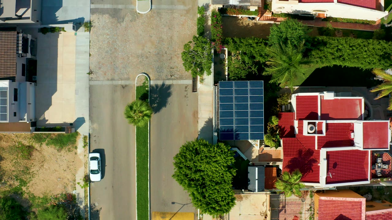 Overhead residential street with a red-roofed house, solar panels, palms, and a white parked car.
