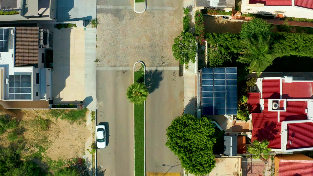 Straight-down view over a San Jose del Cabo residential street with a white car parked along the road, red-roofed homes, rooftop solar panels, palms, dirt ground, and no visible people.