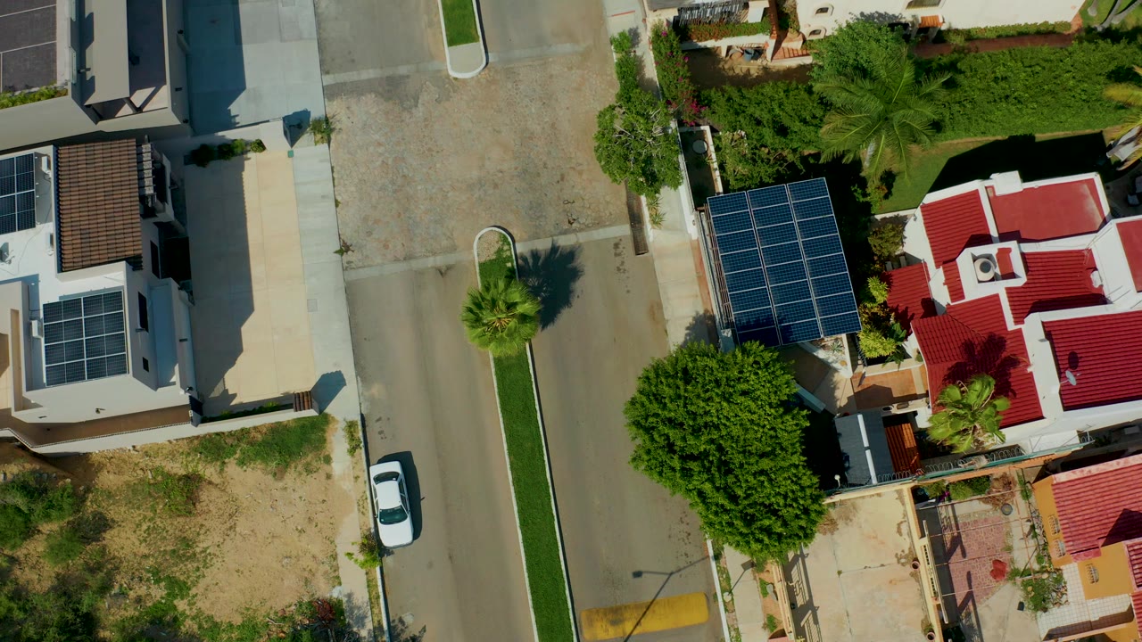 Top-down aerial frame in San Jose del Cabo showing a residential street beside white houses, rooftop solar panels, red roof sections, trees, and a parked white car with no visible people.