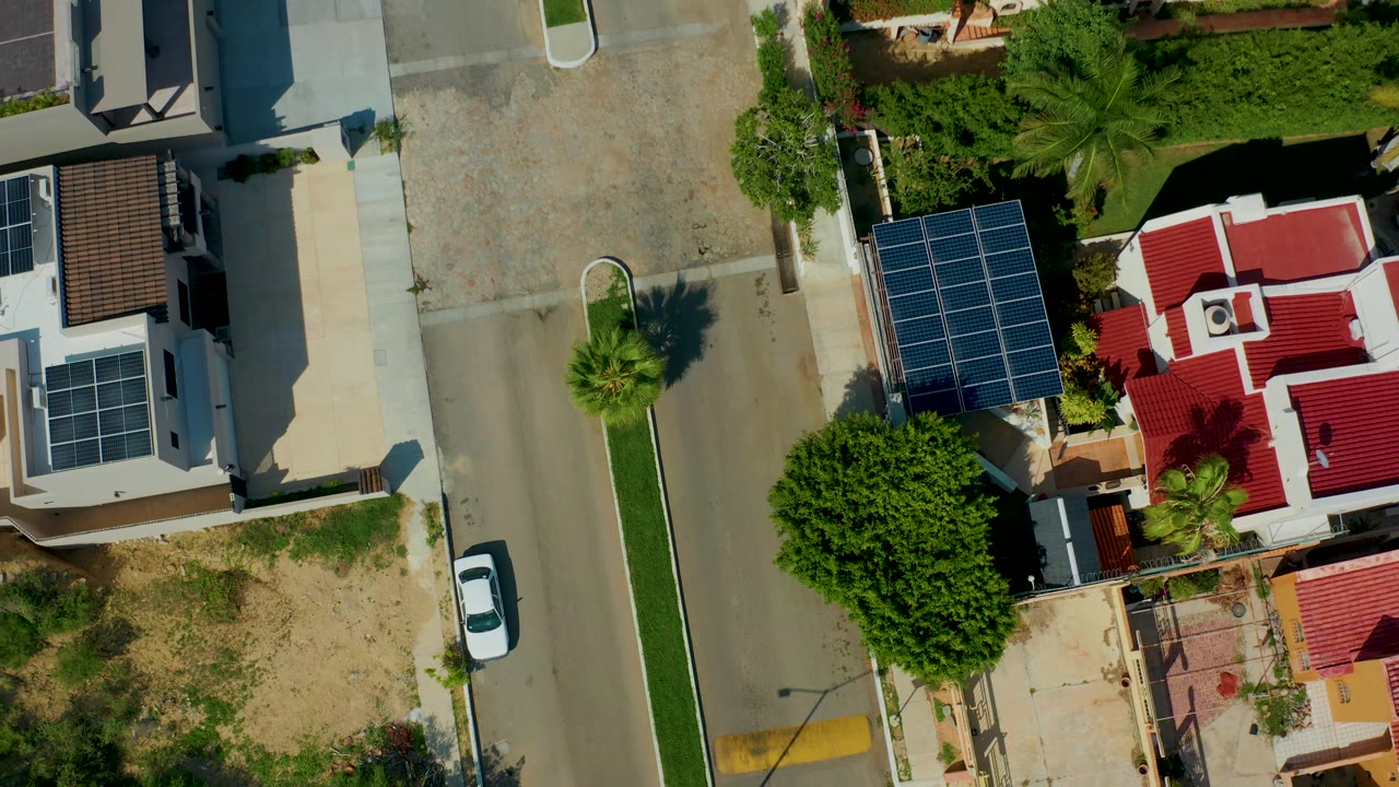 San Jose del Cabo overhead frame showing an empty residential road beside solar-panel rooftops, a red-roofed home, dense green trees, pale driveways, and no visible people.