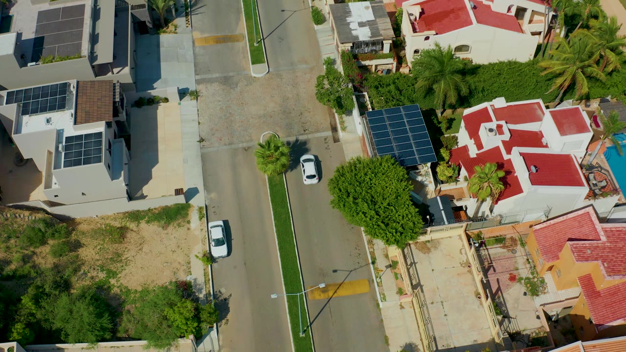 Overhead frame in San Jose Del Cabo showing a white car near the left side of a paved residential street, with red and gray roofs, rooftop solar panels, trees, and no visible people.