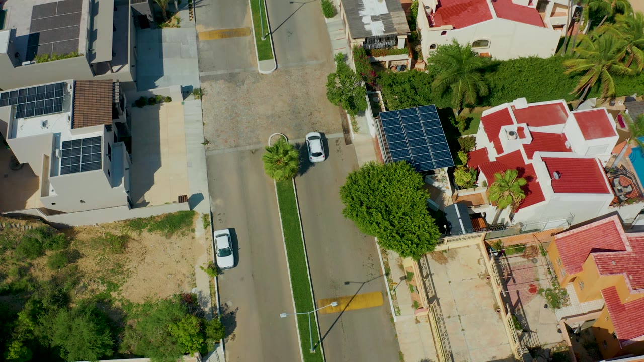 Top-down San Jose Del Cabo frame showing the white car advancing along the residential street, with a red-roofed house, solar-panel arrays, palms, shrubs, and no visible people.
