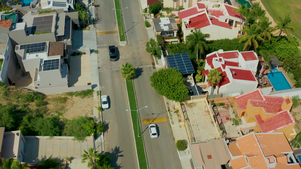 Overhead view of a San Jose del Cabo residential street with solar panels on rooftops, red-roof houses, a white car on the road, a blue pool, palms, and no visible people.