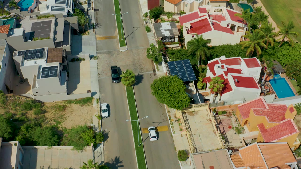 Drone-style overhead view of San Jose del Cabo homes with red and gray roofs, rooftop solar panels, a bright blue pool, a white car, a black car, and no visible people.