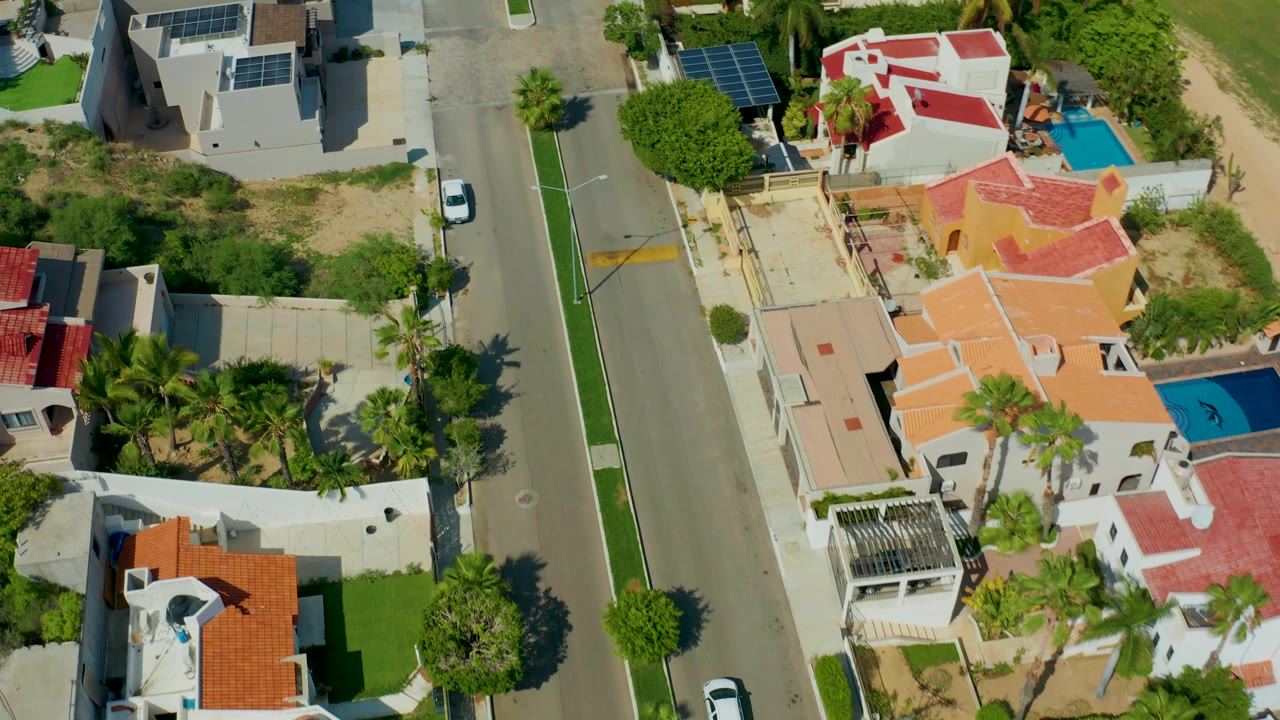 Overhead aerial frame in San Jose del Cabo showing a residential road, walled homes with red and orange roofs, palm trees, solar panels, blue pools, a white car near the road edge, and no visible people.