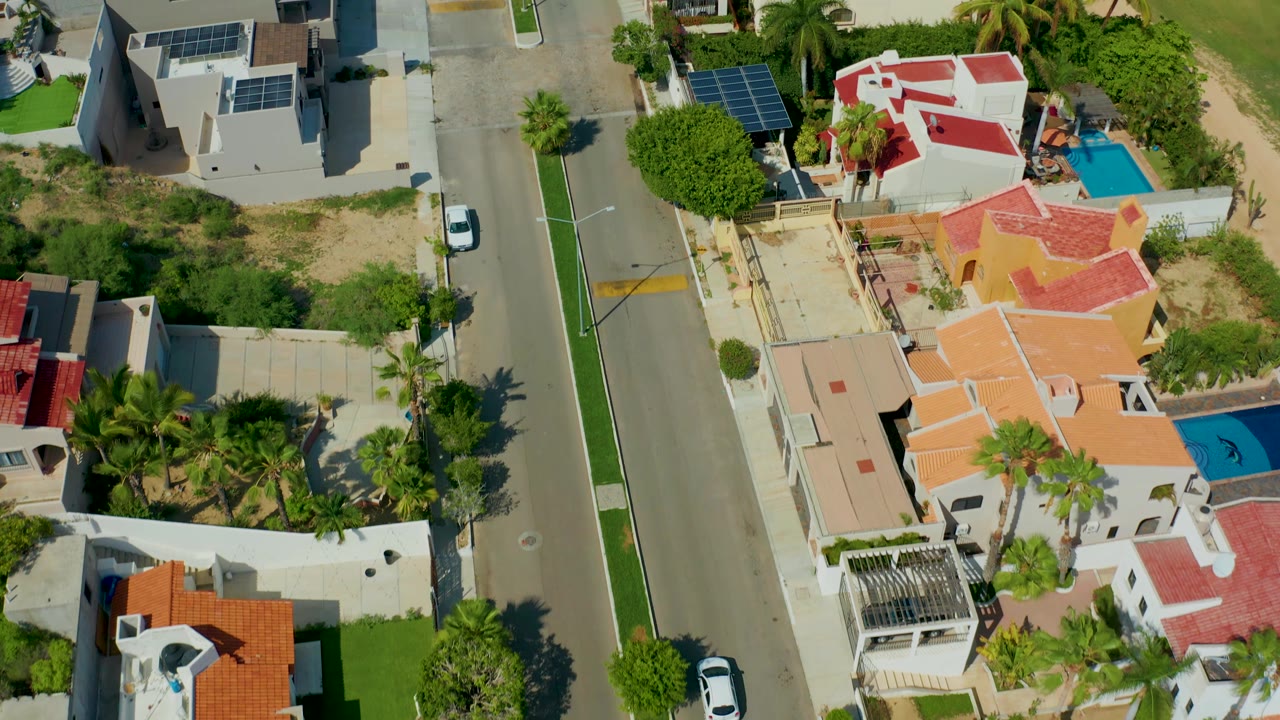 High overhead San Jose del Cabo frame showing the white car near the lower section of the road, a gray home with rooftop solar panels, red-roofed houses, two blue pools, greenery, and no visible people.