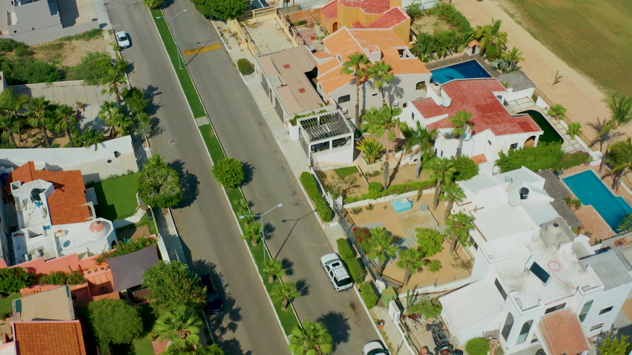 Overhead San Jose del Cabo residential view with a straight street, white and tan houses, red tile roofs, blue pools, palm trees, and no visible people.