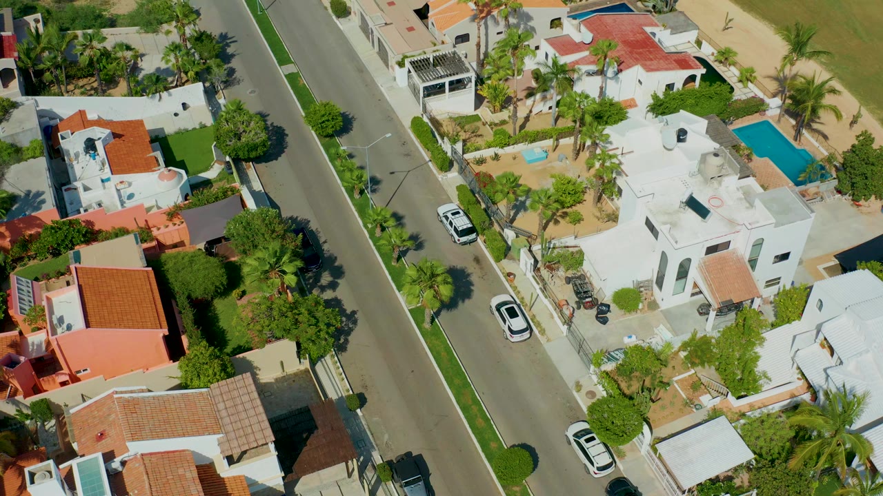 Aerial frame in San Jose Del Cabo showing the road shifted slightly through frame, with white homes, red tile roofs, palm trees, blue pools, and no visible people.