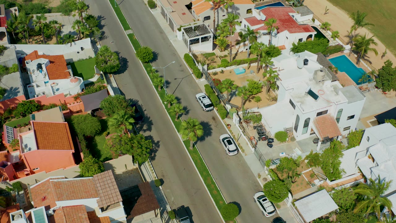 High-angle view of houses, palms, pools, and parked cars along a sunny neighborhood street.
