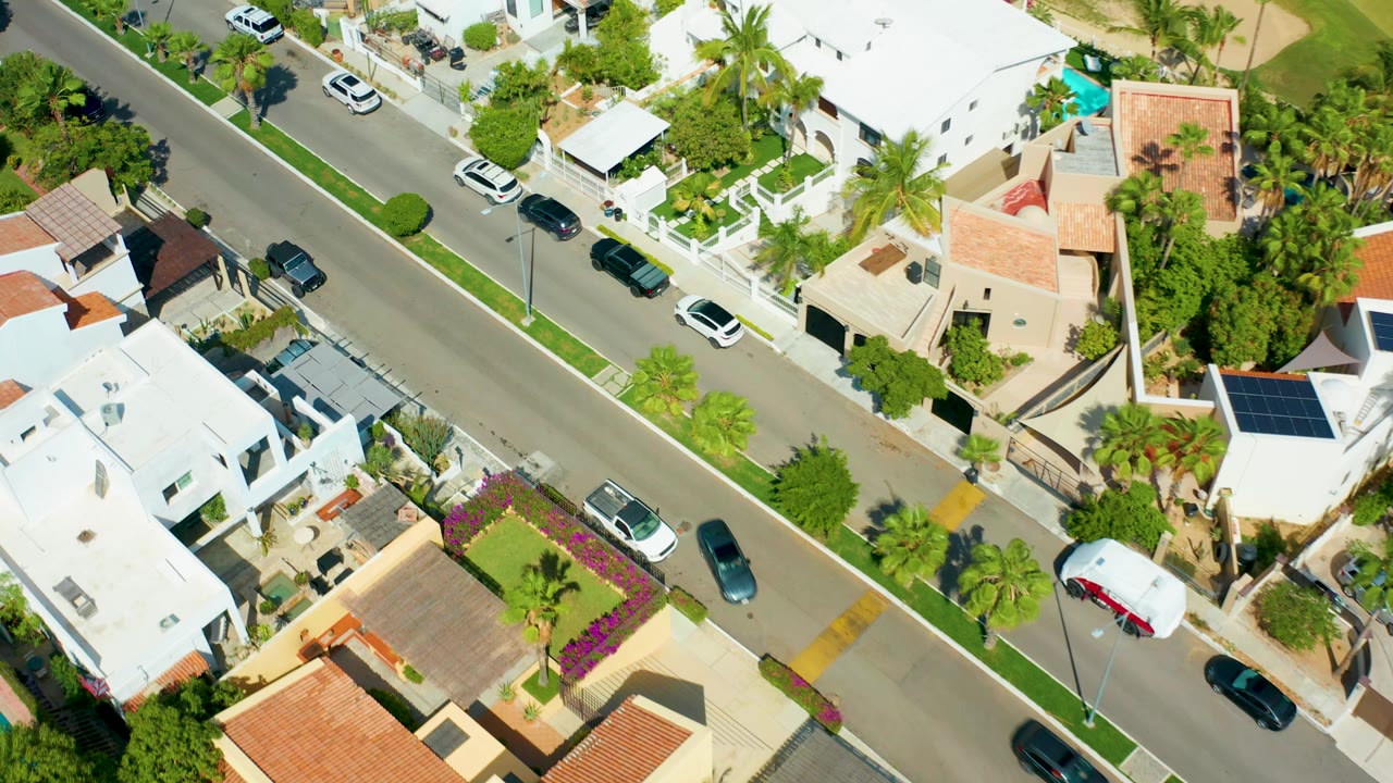 Overhead residential block with a road, cars, palm trees, and white-roofed houses.