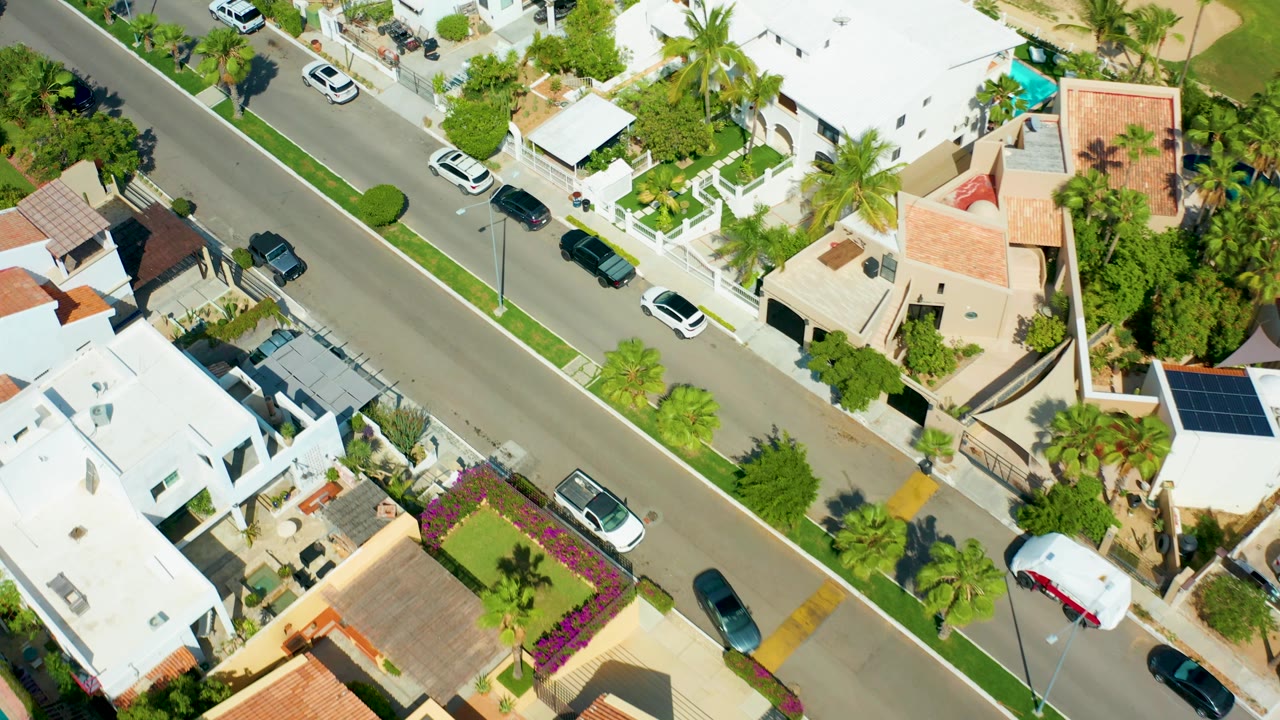 Drone-height view over a San Jose Del Cabo neighborhood where the road angles past villas, curbside cars, palms, trimmed greenery, and a white SUV, with no visible people.