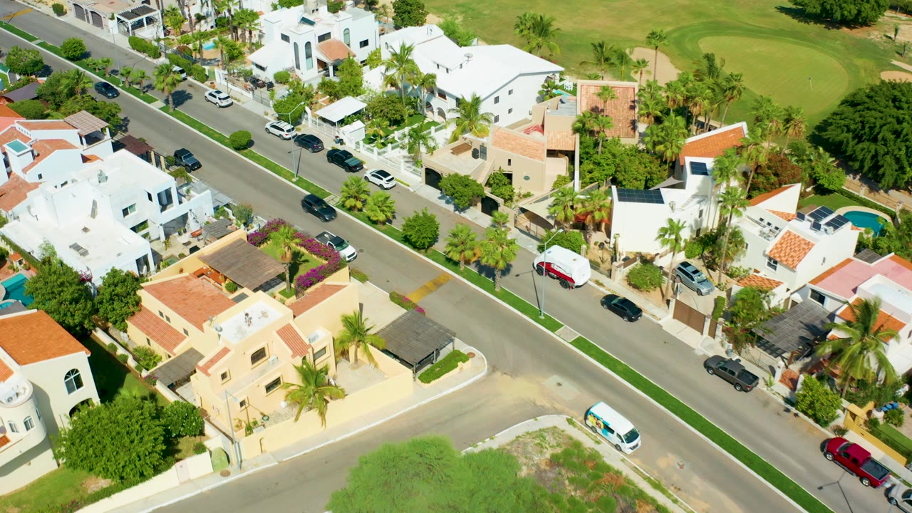 Opening aerial frame over San Jose Del Cabo homes, a diagonal residential road, palms, green lawns, parked cars, and no visible people.