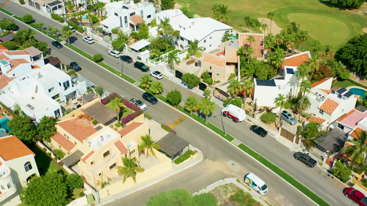 At 0:02, the San Jose Del Cabo aerial view shows cars farther along the residential street, with white and tan homes, palms, lawns, and no visible people.