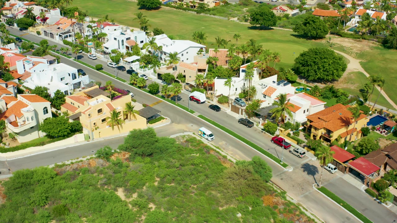 Aerial view in San Jose Del Cabo showing the white van farther along the road, with cream and white houses, red roofs, palms, driveways, parked cars, and no visible people.