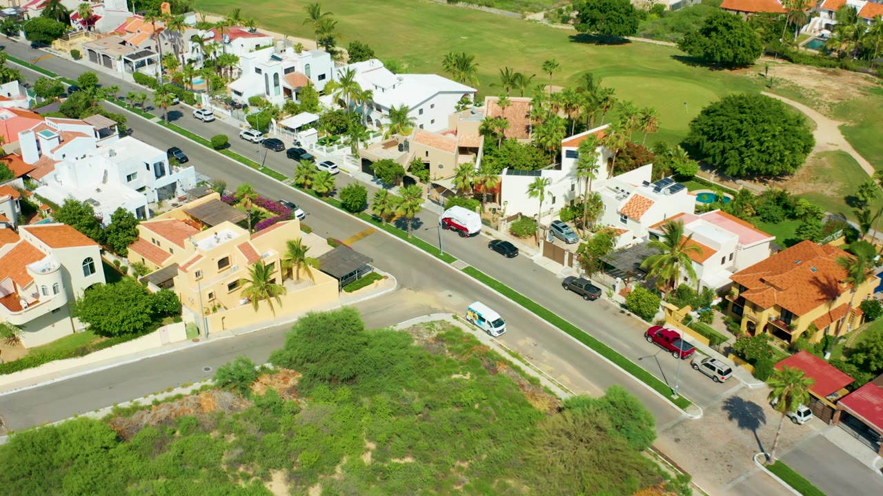 Residential street from above with a white van, parked cars, palms, and open grass.