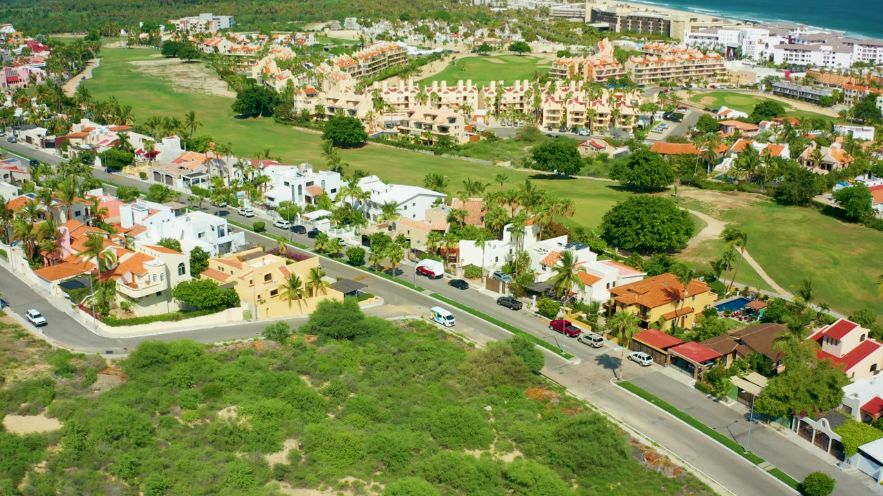Aerial view over San Jose Del Cabo homes with red and white roofs, a curving residential road, parked cars, green open space, scrub vegetation, and no visible people.