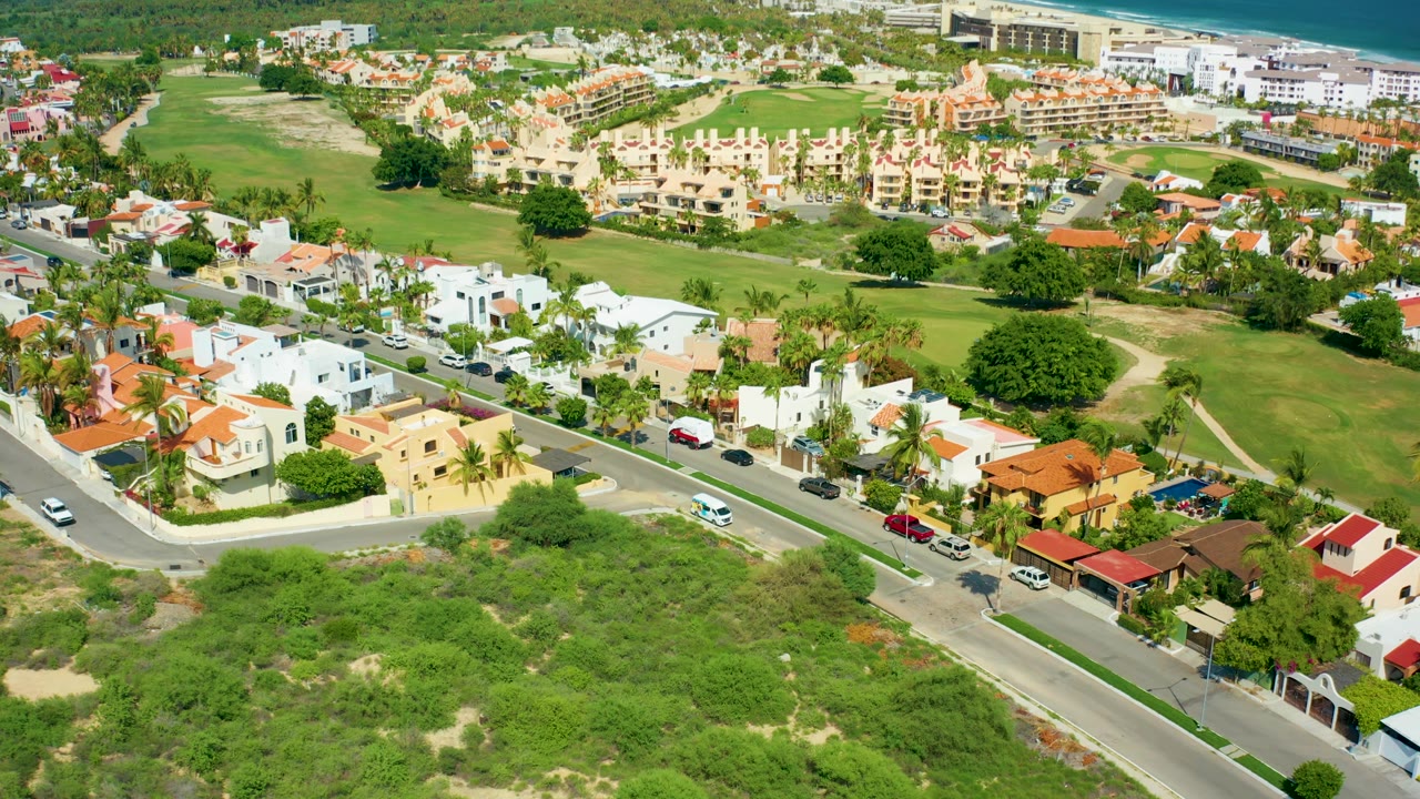 Drone view of a residential street lined with houses and parked cars beside green areas.