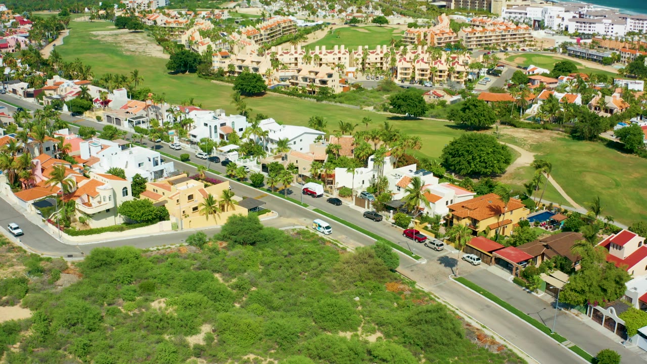 Oblique aerial frame over San Jose Del Cabo villas and homes, with a road crossing the lower scene, several parked cars, palms, open green land, distant buildings, and no visible people.