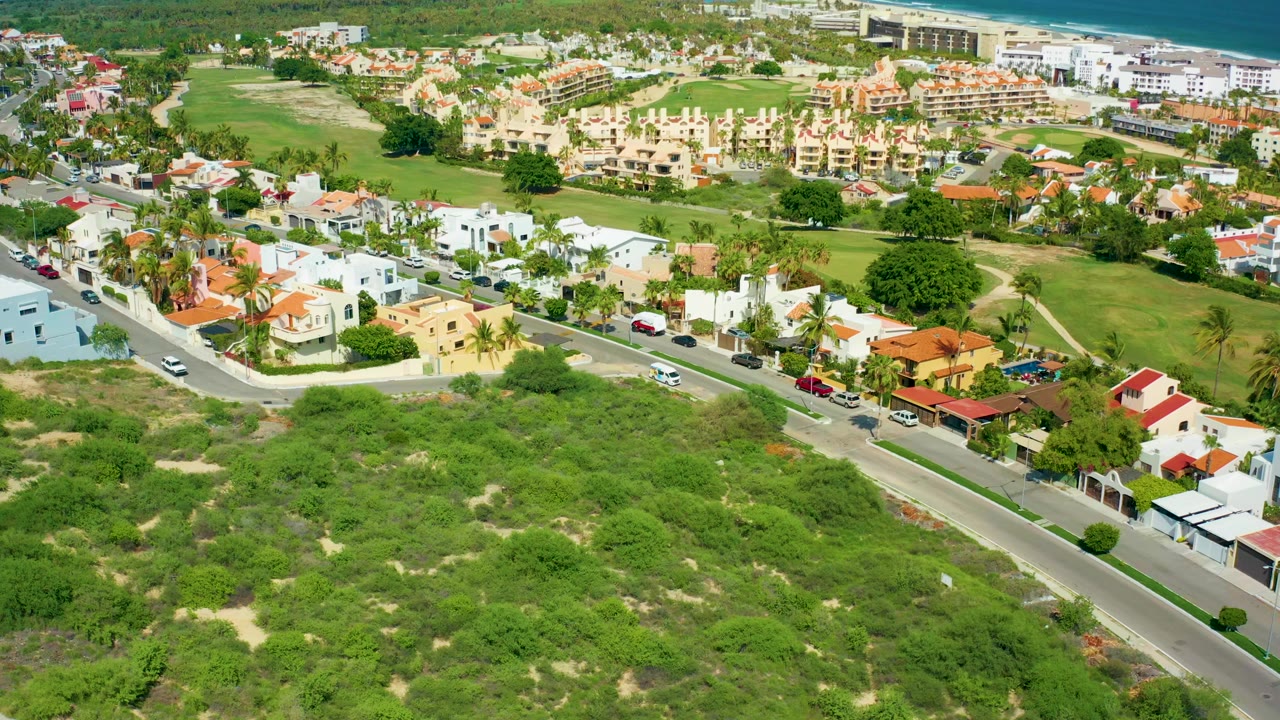 Aerial frame over San Jose Del Cabo showing a paved residential road in the foreground, white and tan houses, green open lots, distant buildings, and a narrow band of ocean, with no visible people.