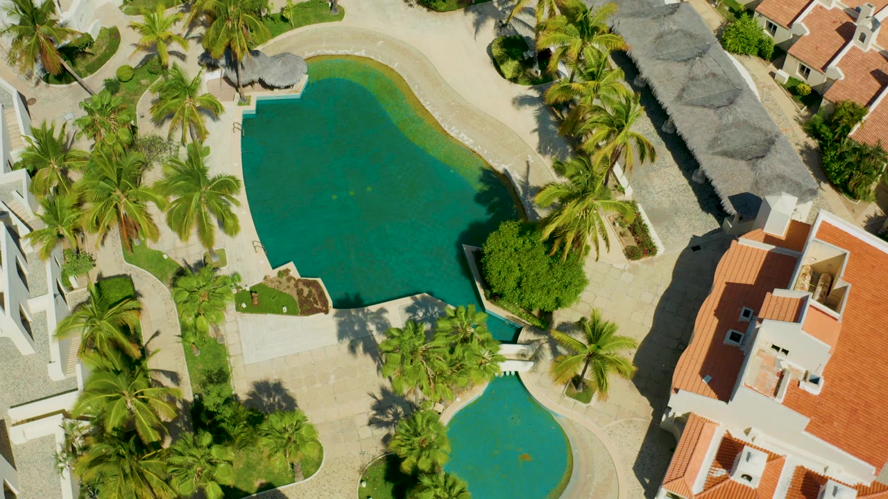 Overhead view in San Jose del Cabo showing green-blue resort pools, palms, pale paths, thatched roof structures, red-tiled buildings, and no visible people at the start of the clip.
