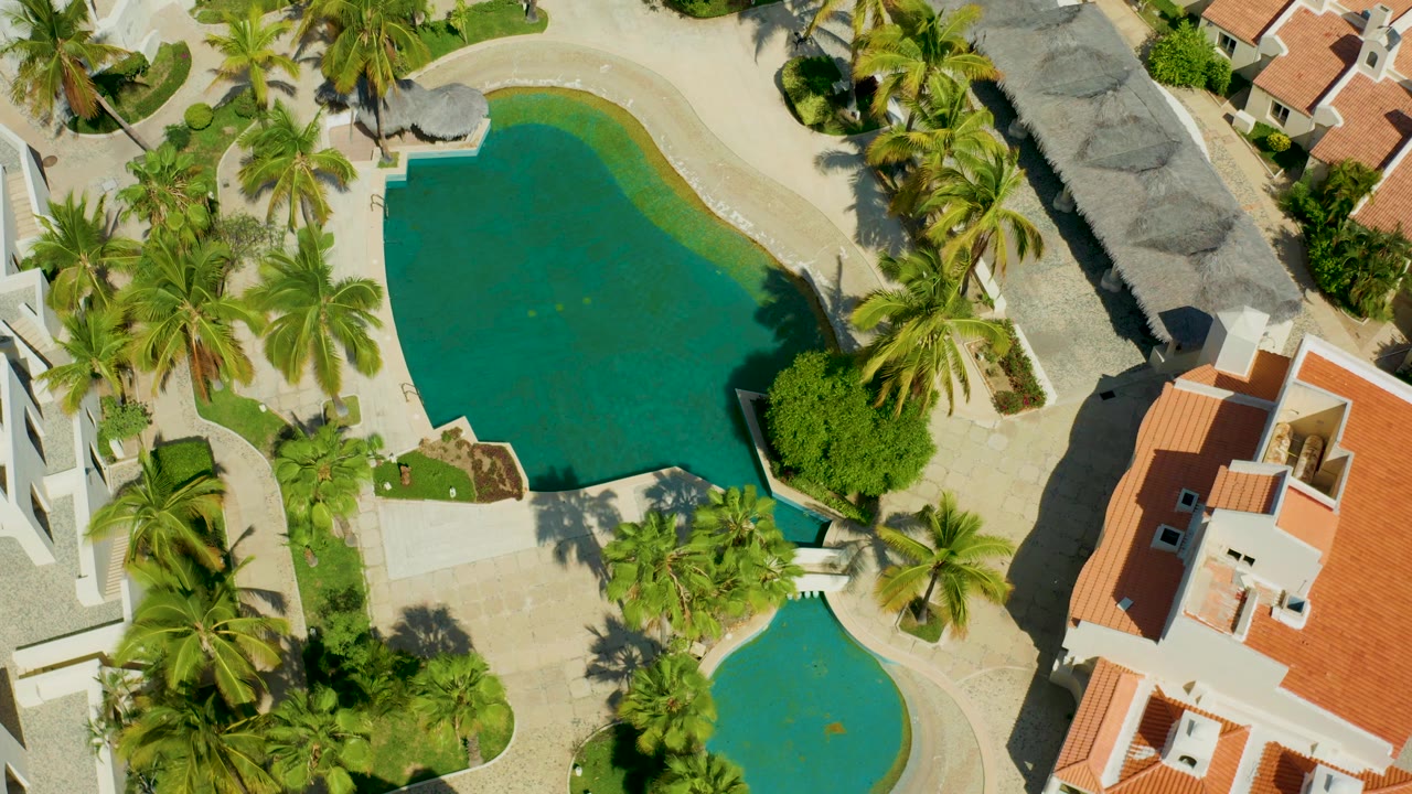 Aerial frame in San Jose del Cabo with two visible pool shapes, clustered palms, pale paving, thatched roof edges, red-tile rooftops, and no visible people.