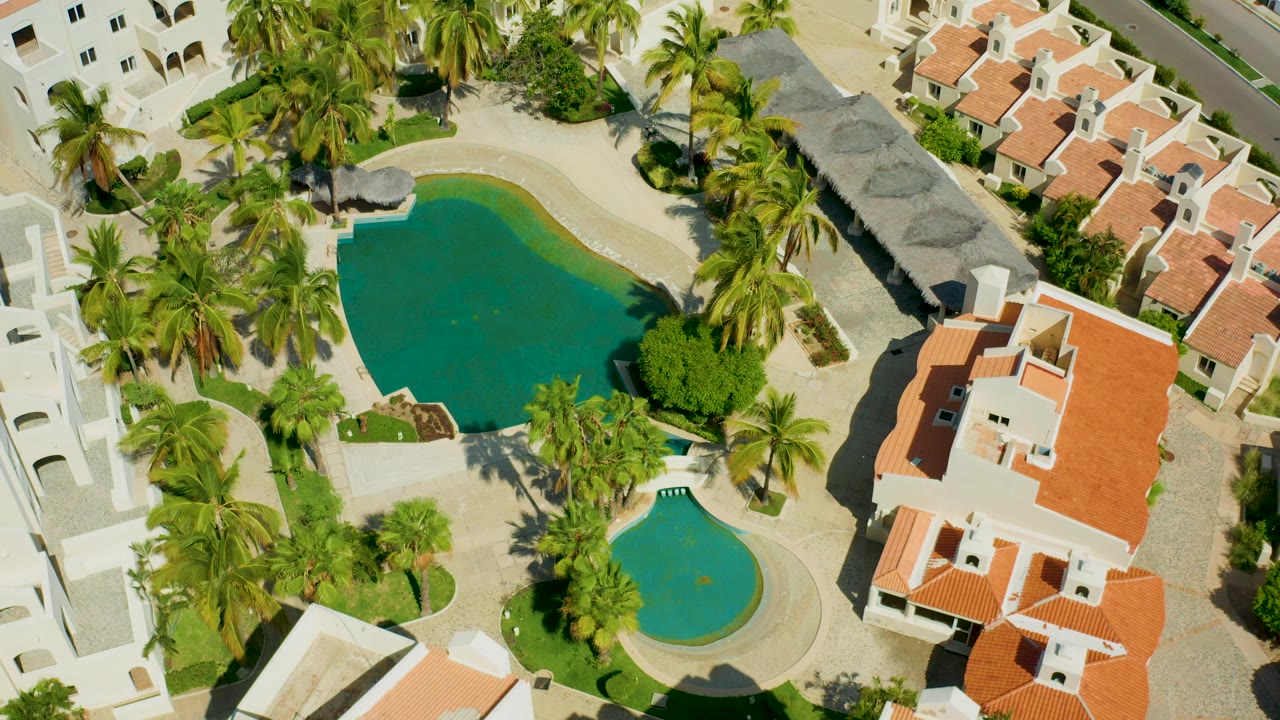 Overhead aerial frame in San Jose del Cabo showing white residential buildings, red tile roofs, two green pools, palms, walkways, and no visible people.