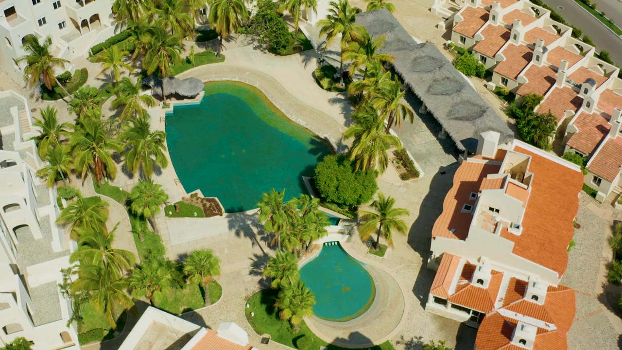 High aerial frame over San Jose del Cabo residences, with two turquoise-green pools, palms casting shadows, white building edges, red roofs, and no visible people.