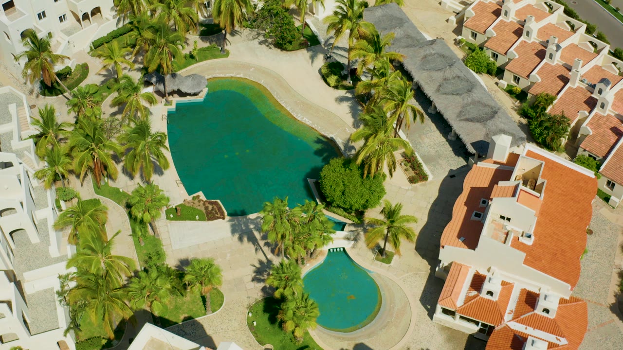 Aerial view closer over the larger pool, palms, paths, and villa roofs.