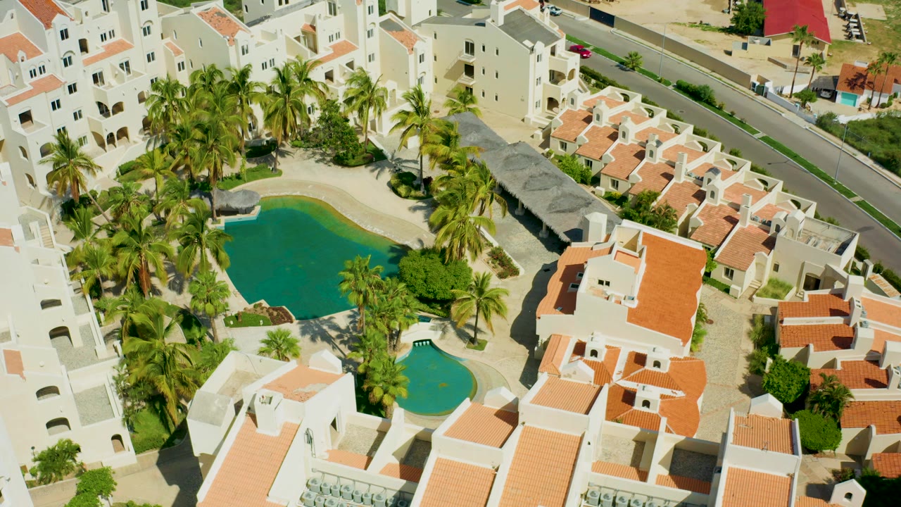 Aerial frame from San Jose del Cabo showing white residential buildings around two blue-green pools, orange tile roofs, palm trees, an adjacent road, and no visible people.